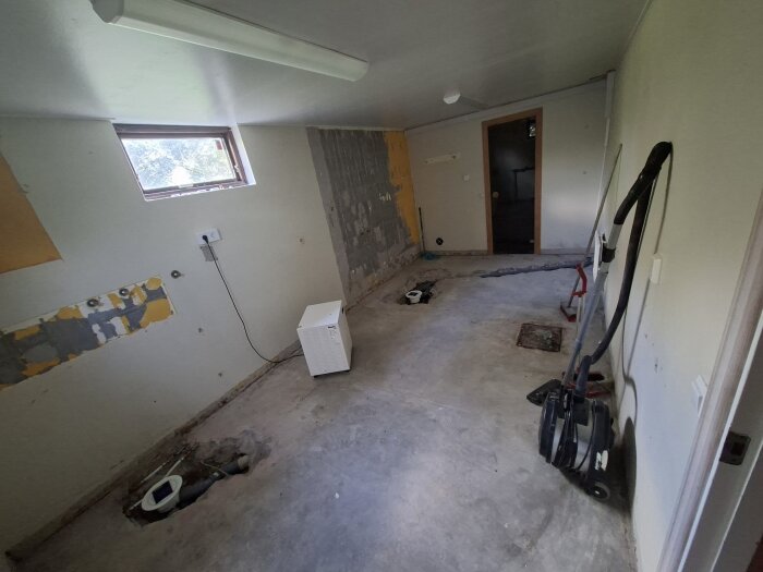 Rum under renovering med utriven golv och väggar, ett fönster, dammsugare och byggverktyg. Väggarna har fläckar av rivet material.