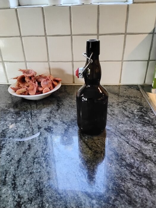 Flaska med patentkork på köksbänk, vitkaklad vägg i bakgrunden, skål med räkor bredvid.