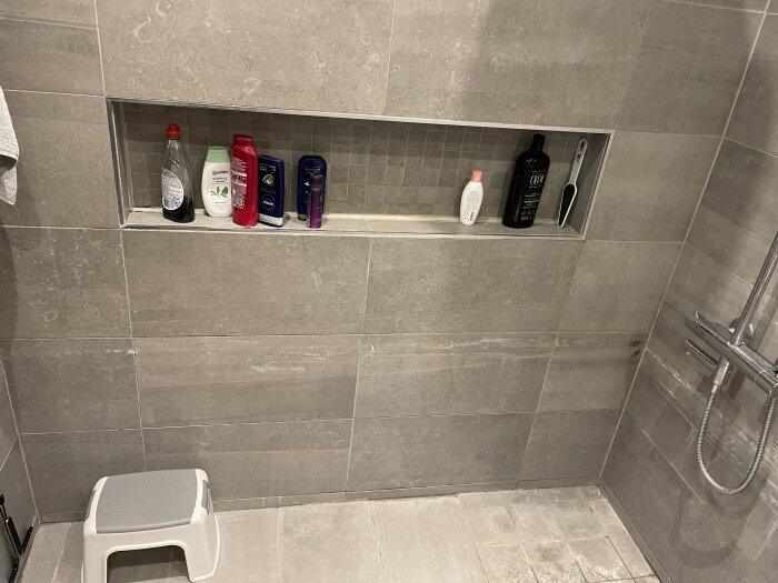 Inbyggd hylla i badrumsvägg med flaskor och tvålar, grå kakelvägg och dusch i bakgrunden.