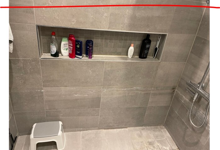 Dusch med grå kakelplattor, en hylla med flaskor och hygienprodukter samt en duschstång med duschmunstycke bredvid.