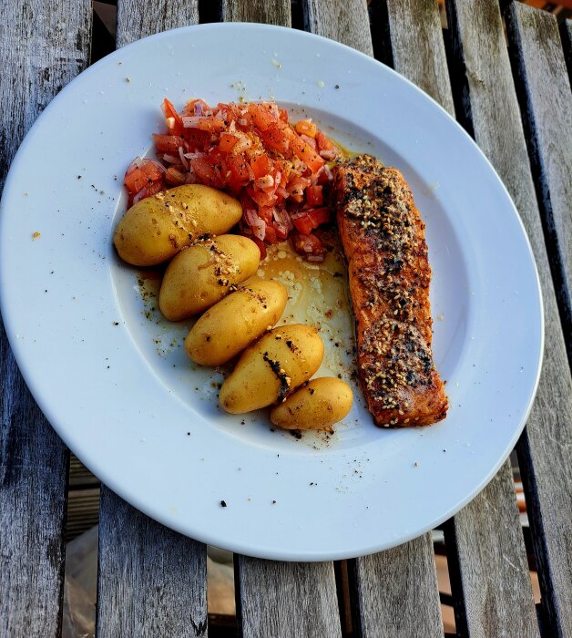 Tallrik med ugnsstekt lax, små potatisar och tomatsallad på en träterrass.