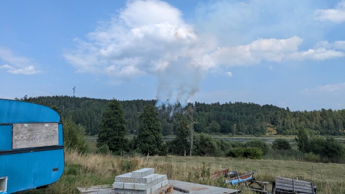 Skogsbrand vid skogsbryn, rökmoln stiger ovan trädtopparna. I förgrunden en blå byggnad, staplade betongblock och verktyg.