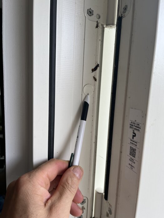 Hand pekar på skruv i karm med penna, visar plats för att sätta extra skruv för att justera sidostycket i en dörr- eller fönsterram.