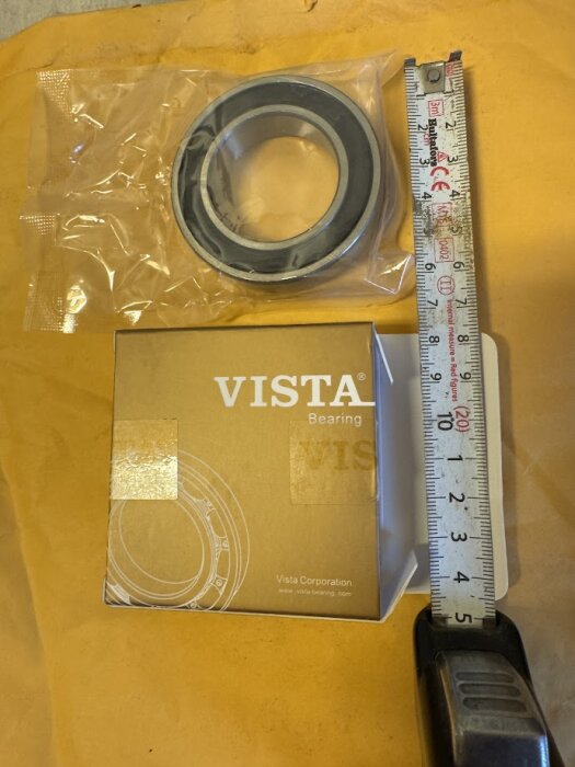 Lager i plastförpackning med måttband och VISTA-förpackning i bakgrunden, används för kraftuttag i gräsklippare.