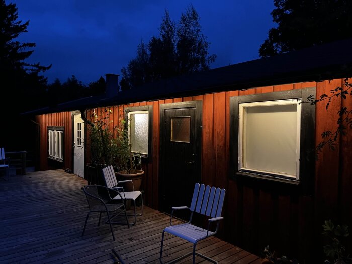 Trähus med orange panel och svarta fönsterramar lyser upp mot en mörk natthimmel. Trätrall med stolar framför byggnaden.