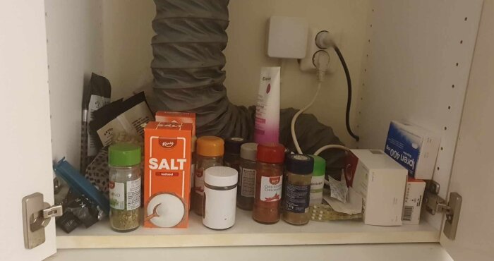 Köksförvaringsskåp med kryddor, saltpaket, och en grå ventilationsslang; sladden tycks vara kopplad till väggen.