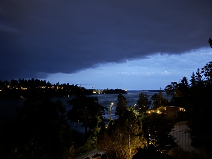Kvällsbild över norra Stockholm, mörka moln som täcker himlen, träd och hus belysta vid vattnet, tecken på annalkande oväder.
