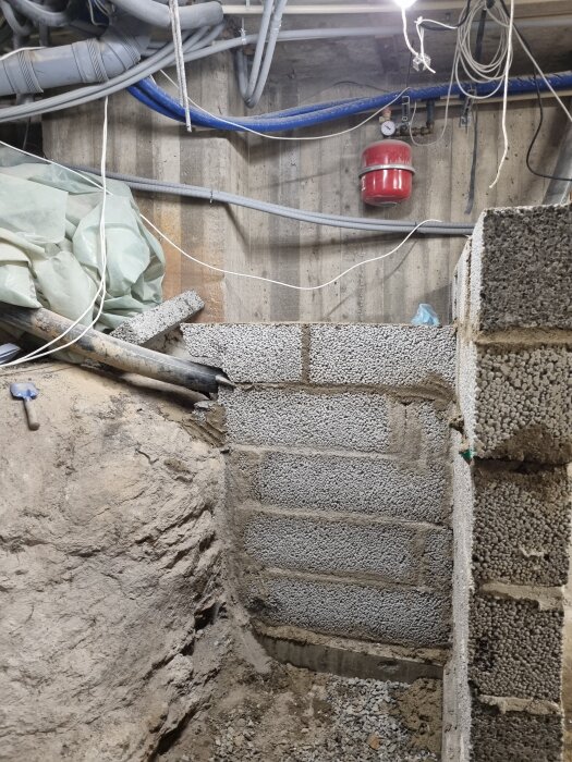 Betongvägg i källare med synliga rör och kablar ovanför. Byggplast och verktyg i bildens vänstra hörn.
