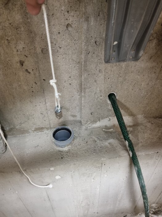Betongvägg med ett hål i, ett grönt rör och en vit snörstump med en bult som hänger ovanför ett röruttag.