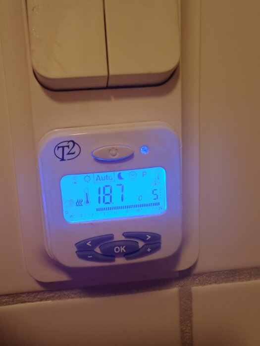 T2-termostat visar 18,7°C med blå bakgrundsbelysning, omgivande väggytor syns.