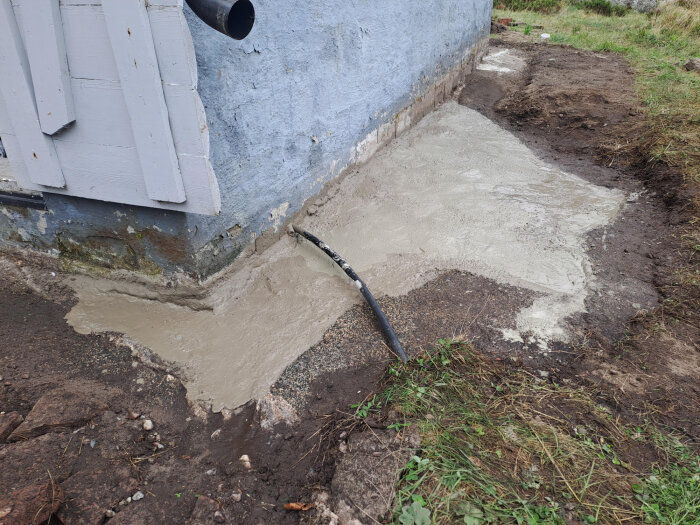 Grund intill husvägg fylld med grus och betong, förberett med dräneringsrör. Markberedning synlig runt omkring, intill blåmålad vägg.