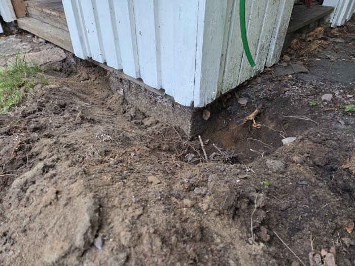 Grävt område vid plintgrund till bod med synlig sand och några rötter, visar arbetet med att förbereda för att lägga grus och laga regel.