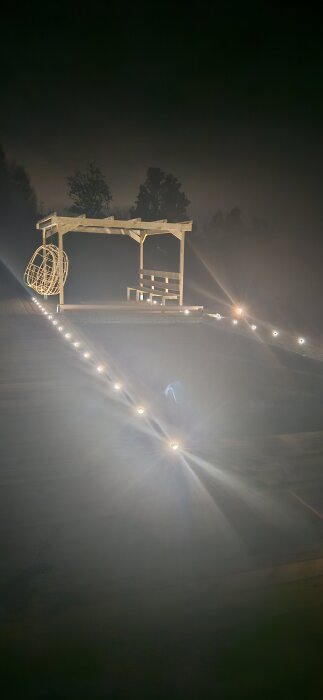 Kvällsbild av en upplyst brygga med sittplats och pergola, omgiven av mörk natur och led-lampor längs bryggan.