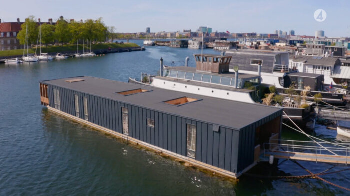 Flytande hus bredvid högre båtar i en hamn, med staden och grönområden i bakgrunden.