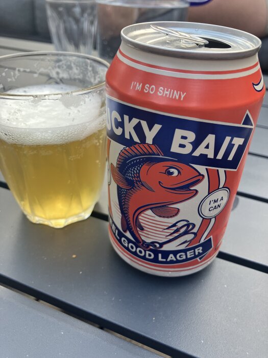 Ölburk med texten "Lucky Bait Feel Good Lager" och bild av en fisk samt ett glas med öl på ett bord utomhus.