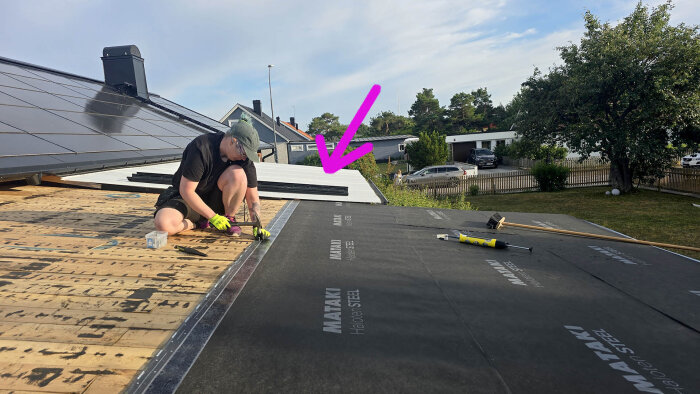 Person monterar uppvik mot husvägg på ett tak med Mataki Holmgaard Steel underlagstäckning, verktyg och material syns på träplankor.