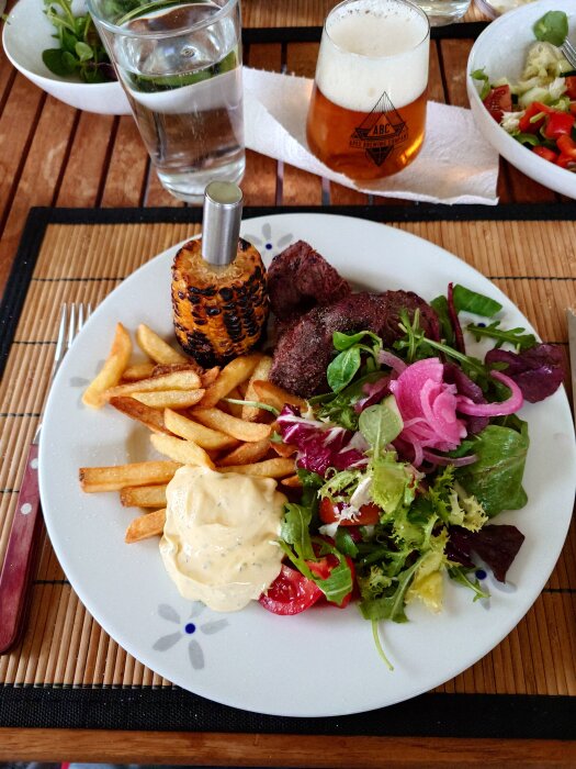 Grillad oxfilé med pommes, bearnaisesÄs, majs och sallad pÄ en tallrik, med vatten och öl vid sidan om, uppdukat pÄ ett trÀbord.