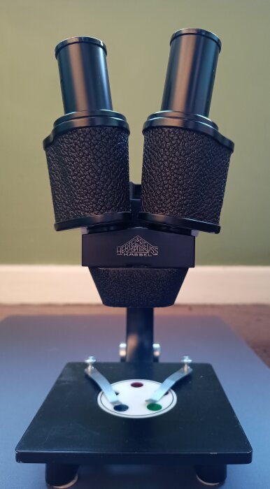 Hertel und Reuss stereomikroskop i närbild, med dubbla okular och svart texturerat grepp, på ett skrivbord mot en grön bakgrund.