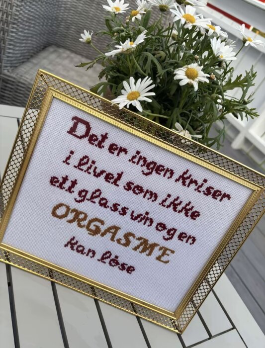 Ramat broderi med humoristiskt budskap på ett bord bredvid prästkragar.
