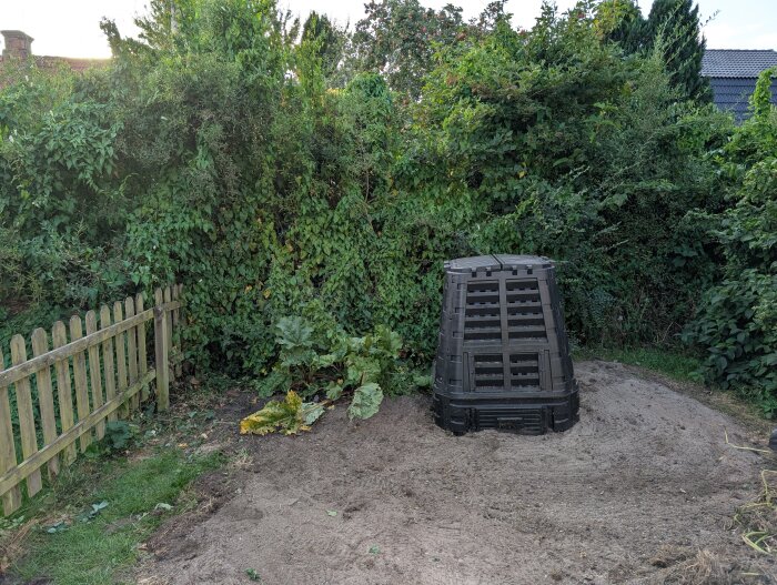 En trädgård med ett staket, tät vegetation och en komposttunna, omgiven av grönska och rensad jord.