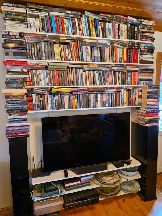 Vägghängd bokhylla med många böcker ovanför en stor TV och högtalare, installerad i en siporexvägg med Elfa-konsolsystem.