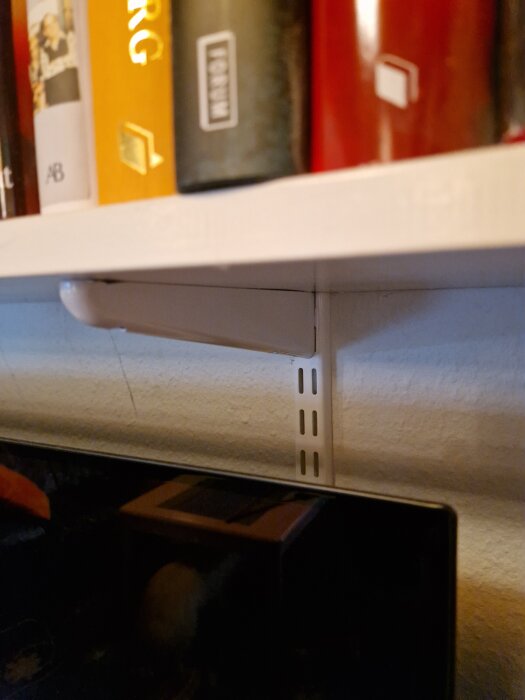 Väggmonterad bokhylla med Elfa-konsolsystem, vit plåtkonsol monterad på lättbetongvägg med böjda pluggar, fylld med böcker.