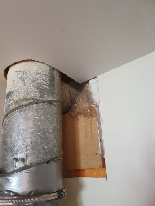 Gammalt ventilationsrör delvis täckt med tejp, passerar genom ett hål i taket. Det ser slitet ut med flagnande yta och skarvar.