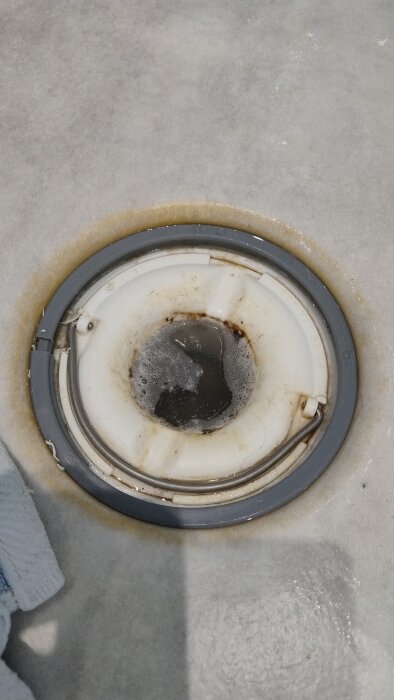 Badrumsgolv med missfärgningar runt en vit golvbrunnslock med tecken på rost eller annan beläggning på locket.