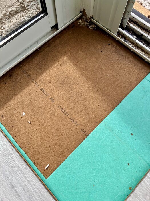 Underlag av masonit i uterummets hörn med fuktfläck och mindre hål, delvis täckt av grön isolering.