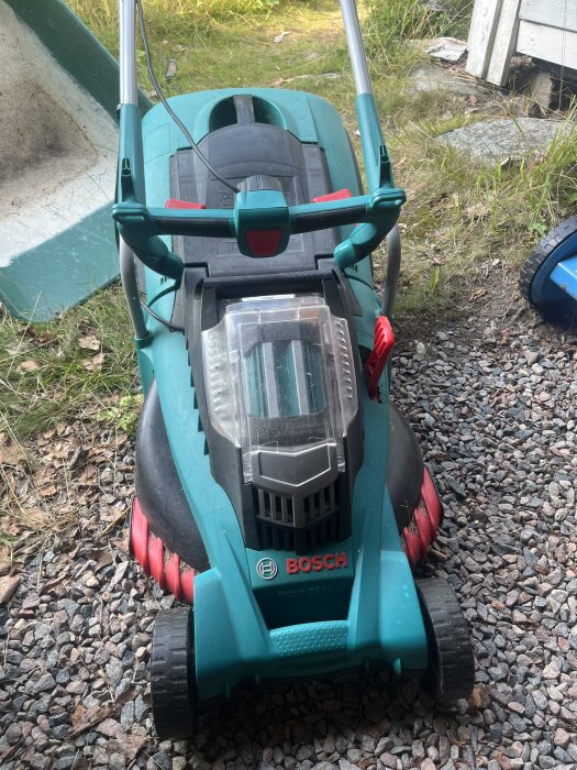 Äldre Bosch Rotak elgräsklippare på grusmark, med synligt batterifack och handtag, omgiven av gräs och stenar.
