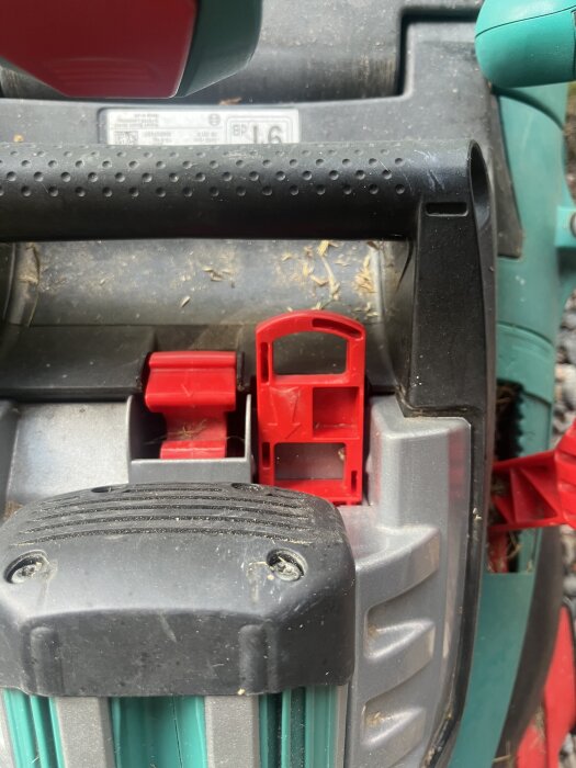Äldre Bosch Rotak elgräsklippare med batterilucka och röd nyckel, synlig del av handtag och slitage samt gräsrester runtom.