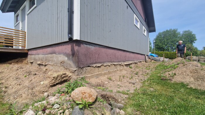 Husets sockel och grund avtäckt med sandig jord runtom, en man står i bakgrunden och inspekterar området.