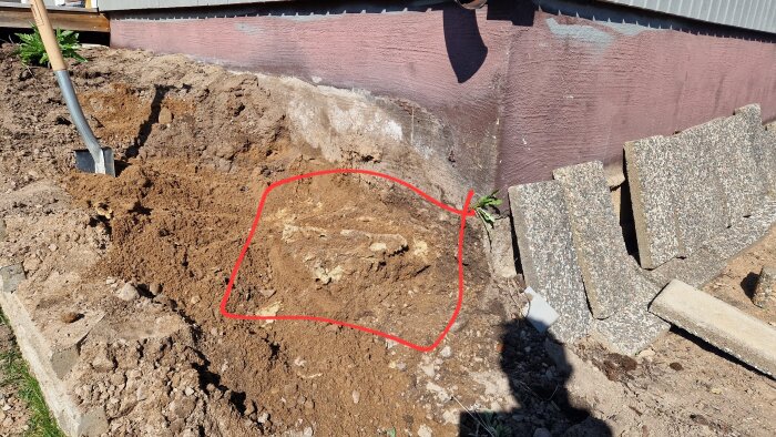 Upptagen markyta vid husgrund, med synliga markskivor och borttagen rabatter, samt stenhögar vid sidan.