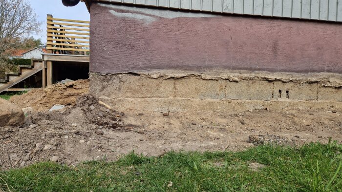 Husgrund med synliga markskivor och jord borttagen längs väggen; verandan synlig i bakgrunden, förberett för ny puts och skydd mot tjäle.