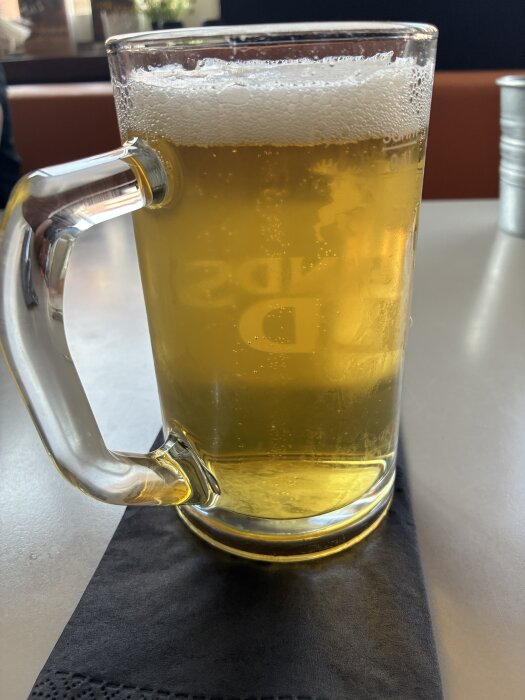 En klar öl i ett glas med skum, placerad på ett svart underlägg på ett bord.