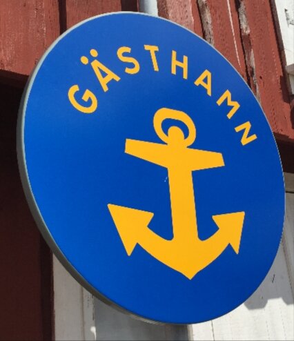 Blå skylt med gul ankare och texten "Gästhamn" mot en röd trävägg.