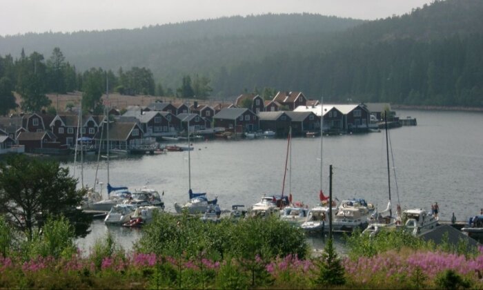 En småbåtshamn med segelbåtar framför en rad röda stugor vid en sjö och skogsklädda berg i bakgrunden.