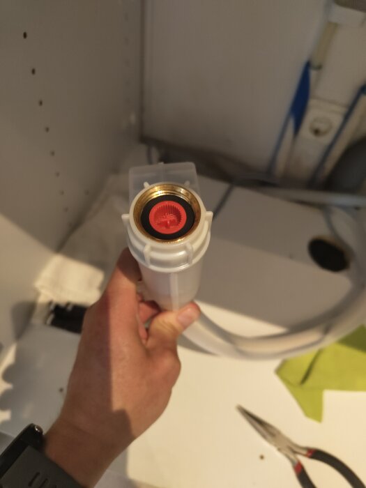 En hand håller en vit diskmaskinslang med en röd insats vid gängan i ett kök; frågeställning om vattenanslutning.