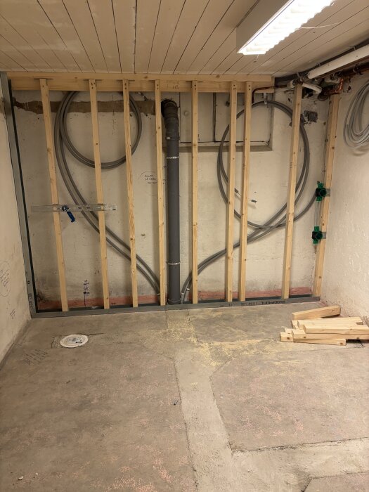 Vägg i byggprocess, uppreglade träreglar och synliga rörledningar. Staplade träbitar på golvet.