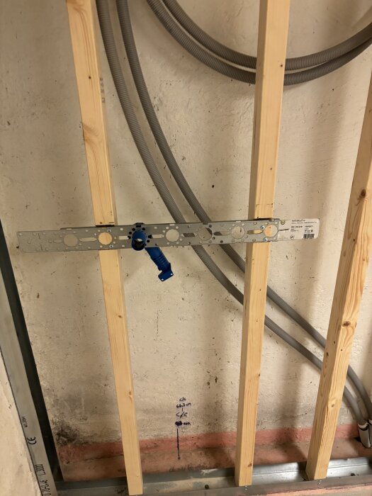 Vägg under konstruktion med träreglar, elrör och en metallplatta med infästning för elinstallation.