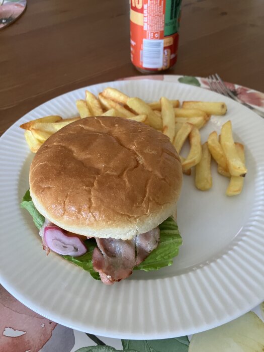 Hamburgare med bacon, sallad och picklad rödlök på tallrik med pommes frites och läskburk i bakgrunden.