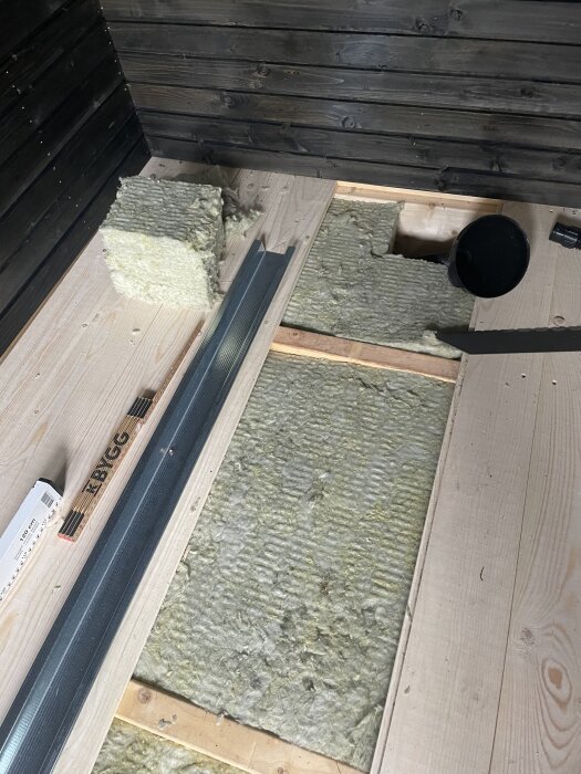 Bastubygge med golv under konstruktion, isoleringsmaterial och grått metallskena synliga. Svarta träväggar i bakgrunden.