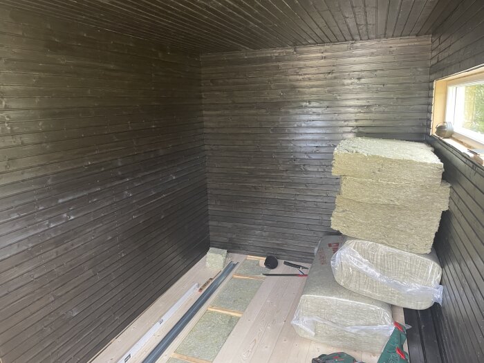 Svartmålat basturum med staplade isoleringsskivor och oisolerat golv under installation. Fönster på ena väggen släpper in ljus.