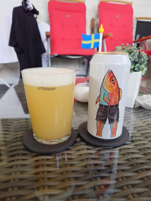 Ett glas med öl från Fermenterarna bredvid en burk dekorerad med en tecknad fisk i shorts på ett bord med svensk flagga i bakgrunden.