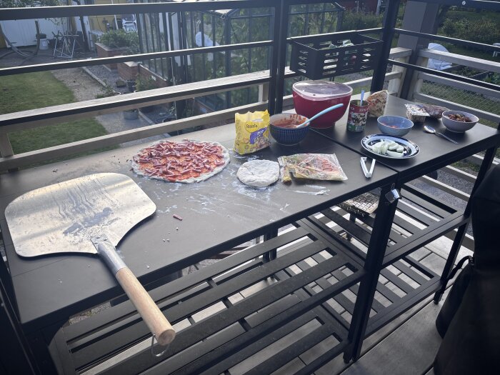 Bord utomhus med pizzatillbehör: tomatsås, deg, paprika och ost bredvid en pizzaspade, redo för pizzabak.