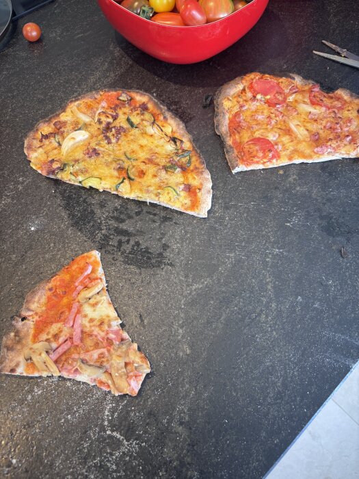 Tre olika pizzabitar med varierande pålägg ligger på en mörk bänk; en röd skål med tomater syns i bakgrunden.