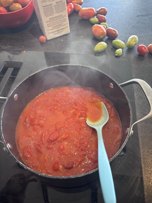 Kasrull med kokande tomatsås, sked i blå plast. Färska tomater och förpackning i bakgrunden på svart köksbänk.
