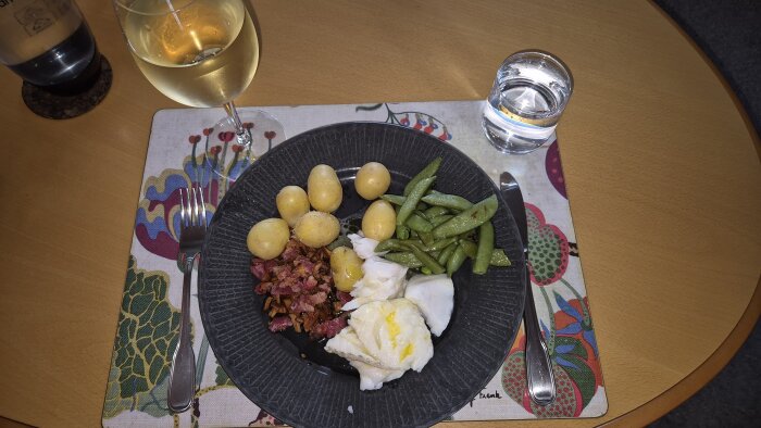 Kokt Skrei med smörstekta kantareller, bacon, sockerärtor och färskpotatis på tallrik. Tillbehör: glas Riesling och vatten på bordet.