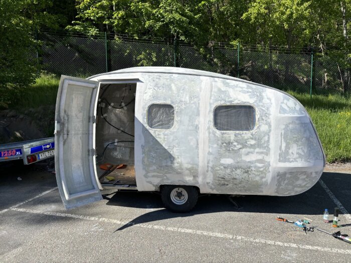 En husvagn under renovering, med öppen dörr och omålad yta, står på parkeringsplats; verktyg och material ligger spridda på marken.