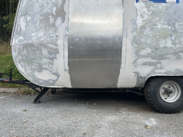 Kaross av husvagn med spackel och synliga aluminiumplåtar, delvis reparerad för sprickor, stående på en asfalterad yta med ett hjul synligt.